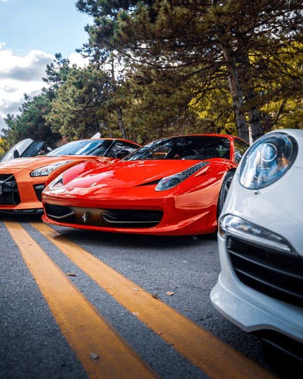 Red Ferrari 458 Italia low front three-quarter on a tree-lined road, flanked by a white Porsche 911 and an orange Nissan GT-R