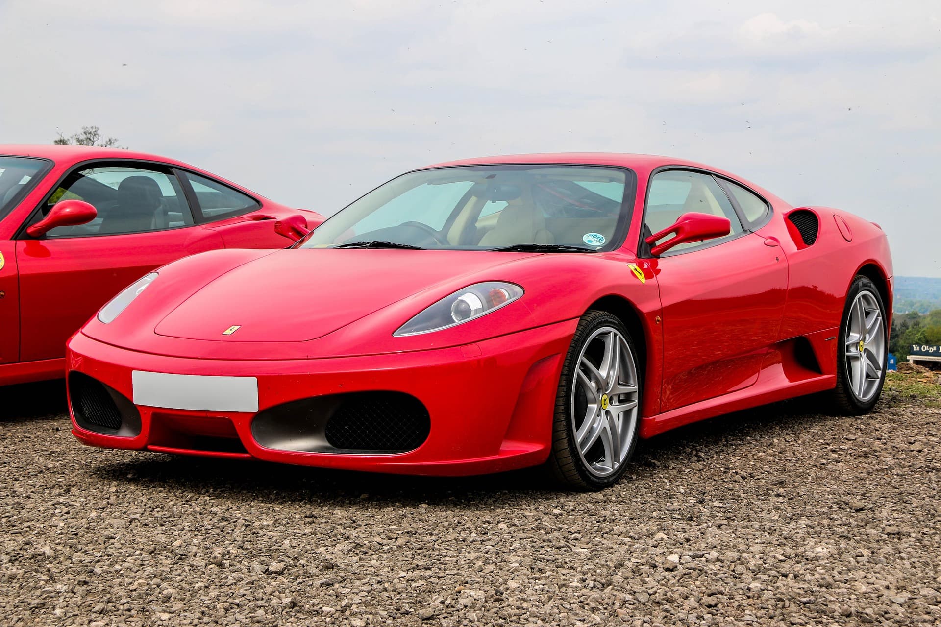 Red Ferrari F430 parked on gravel beside another Ferrari under an overcast sky