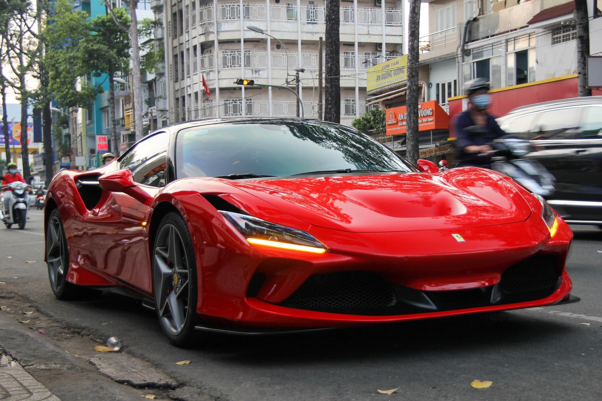 Red Ferrari F8 Tributo with indicators lit, driving along a busy tree-lined city street