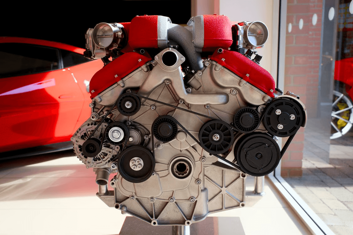 Ferrari flat-plane V8 engine mounted on a display stand, showing red crackle-finish intake plenums and the accessory belt drive