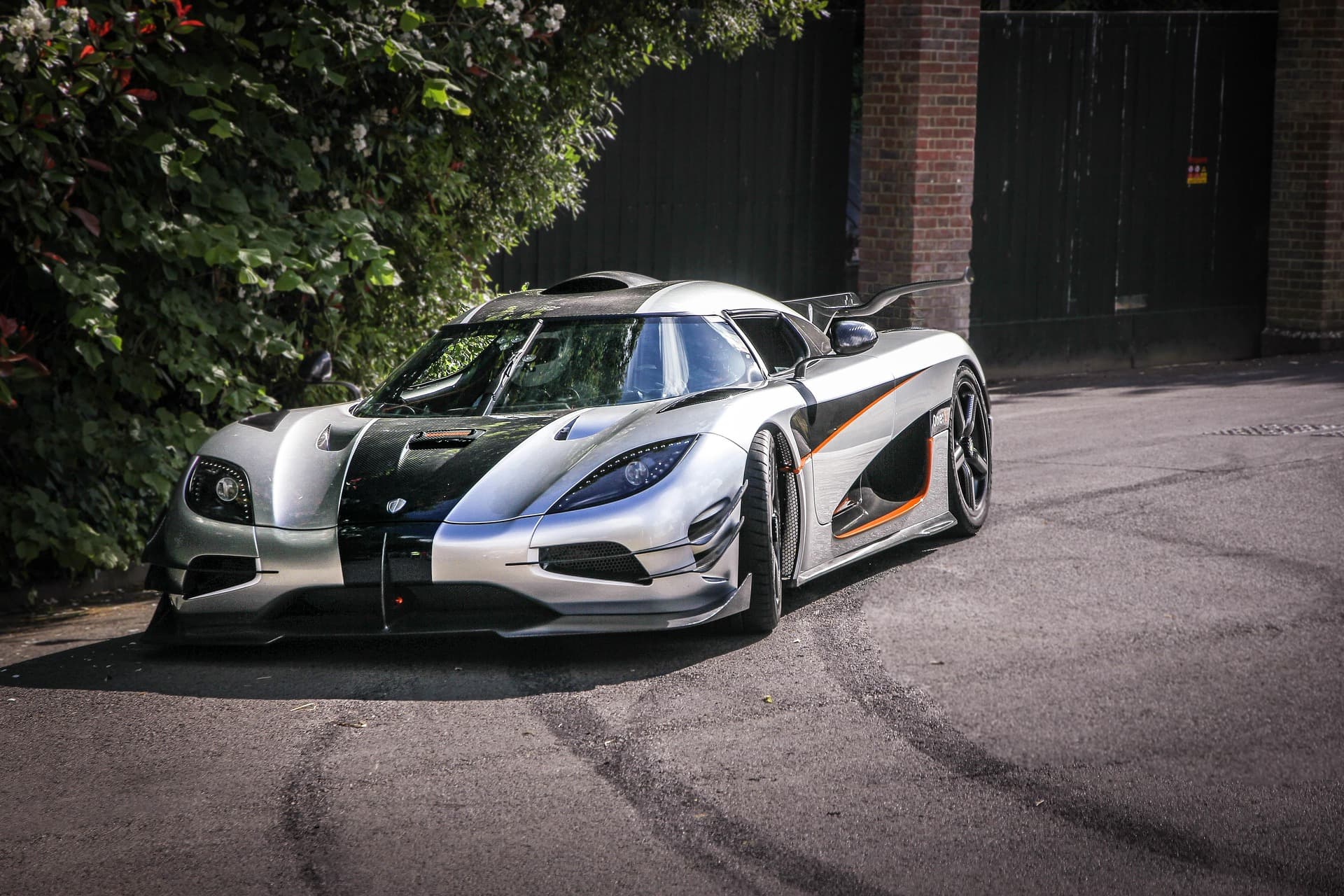 Silver-and-carbon Koenigsegg One:1 with orange accents parked on a driveway beside a brick gateway