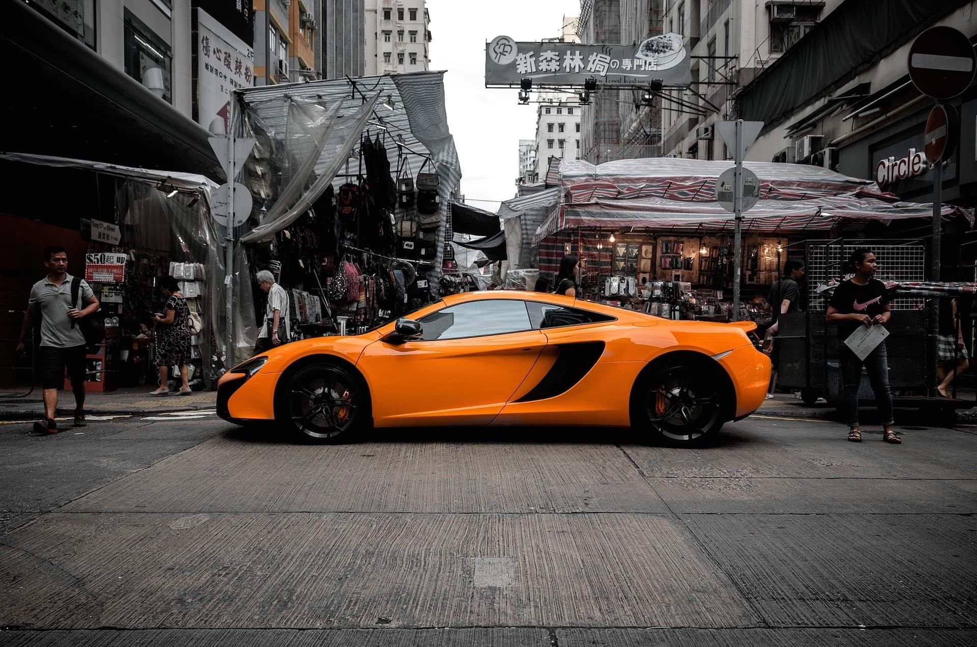 Orange McLaren 650S in side profile crossing a Hong Kong market street lined with stalls