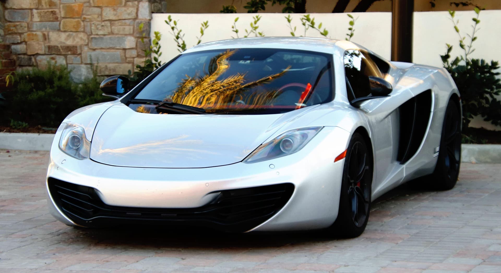 Silver McLaren MP4-12C parked on brick paving in front of a stone wall, palm fronds reflected in the windscreen